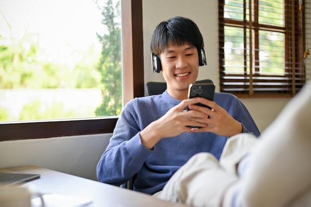 Knappe Aziatische man ontspannen in zijn kantoor aan huis zijn benen op tafel zetten met behulp van telefoon