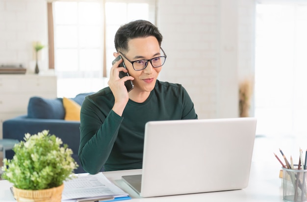 Knappe Aziatische zakenman zittend aan tafel met laptop en praten op smartphone in kantoor aan huis.