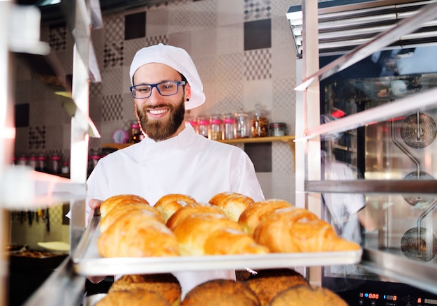 Knappe bakker in witte uniforme houden in zijn handen een dienblad vol met vers gebakken croissants tegen het oppervlak van een bakkerij