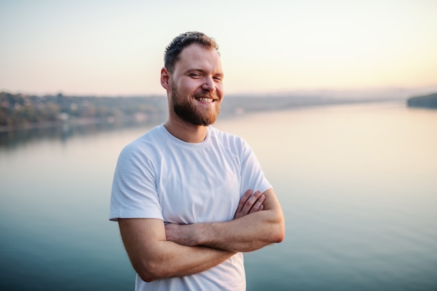 Knappe bebaarde blonde blanke man die op klif met armen gekruist door de rivier.