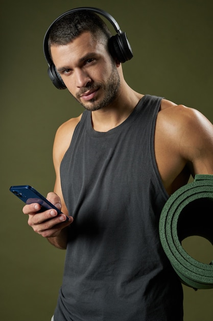 Knappe bebaarde fitnesstrainer in koptelefoon met fitness mat in de hand geïsoleerd op groene achtergrond