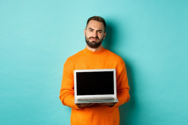 Knappe bebaarde man in oranje trui laptop scherm tonen, promo demonstreren, grimassen teleurgesteld en boos staande over lichtblauwe achtergrond.