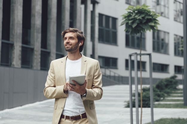 Knappe bebaarde man in stijlvol beige pak met behulp van digitale tablet terwijl je in de buurt van donker modern gebouw