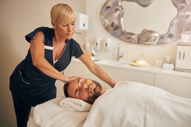 Knappe bebaarde man liggend op massagetafel onder witte deken terwijl hij therapeutische massage krijgt in de spa salon