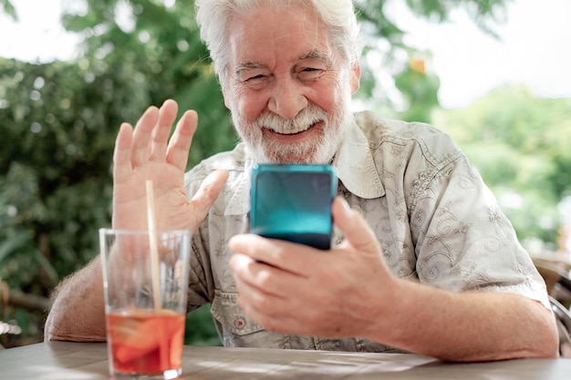 Knappe bebaarde man uit de jaren 70 die buiten aan cafétafel zit en videochat via de mobiele telefoon