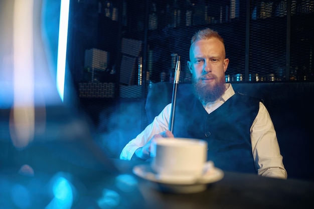 Knappe bebaarde man zittend aan tafel en waterpijp roken terwijl hij koffie drinkt in de bar. Selectieve focus