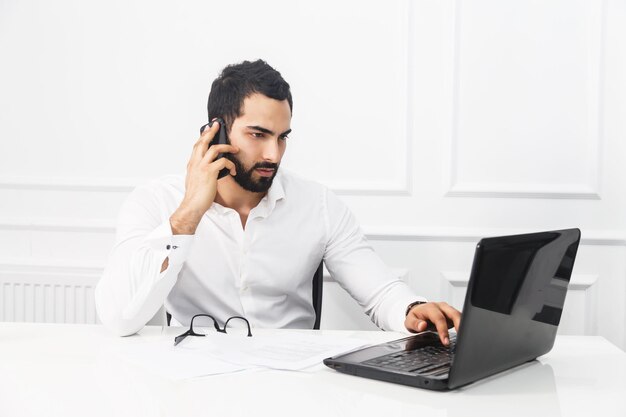 Knappe bebaarde zakenman draagt een wit overhemd op laptop en praat via de telefoon in een wit kantoorinterieur