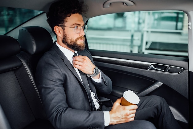 Knappe bebaarde zakenman zit met koffie om op de achterbank van de nieuwe auto te gaan
