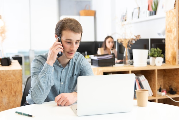 Knappe blanke jonge man praten op zijn mobiele telefoon voor zijn laptop op kantoor.