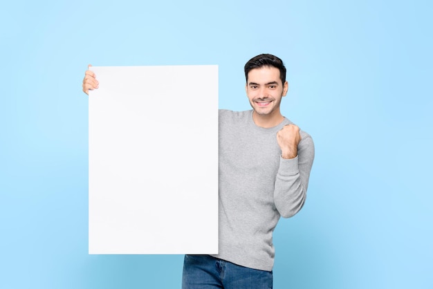 Knappe blanke man die een leeg Witboekbord met een vuist op een lichtblauwe geïsoleerde achtergrond toont