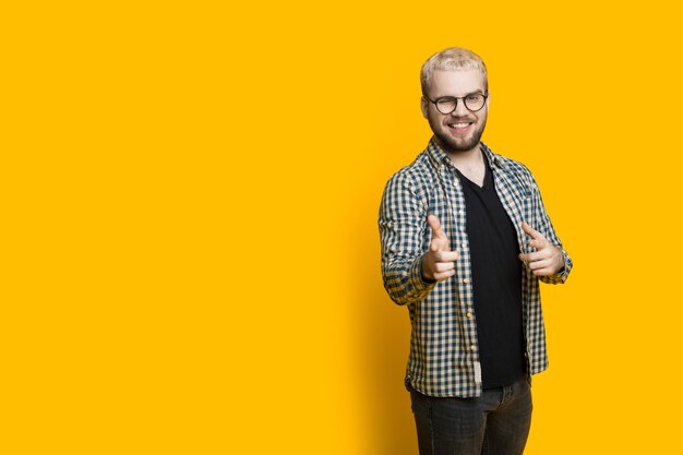 Knappe blanke man naar voren wijzend met wijsvinger dragen van een bril en blond haar poseren op een gele muur met vrije ruimte