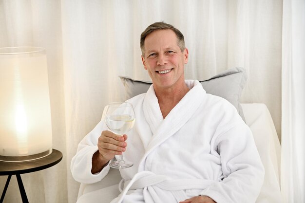 Knappe blanke man van middelbare leeftijd in witte badstof badjas houdt een drinkglas met water vast, glimlacht naar de camera kijkend, genietend van zijn vrije dag in een privémassageruimte van een luxe wellness-spacentrum