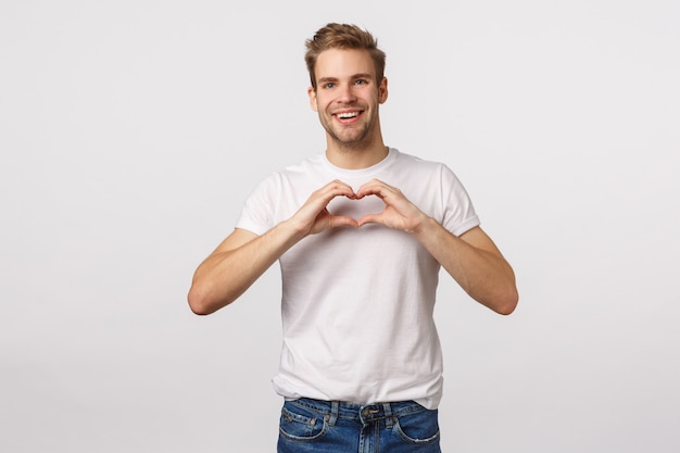 Knappe blonde kerel met blauwe ogen en witte T-shirt die hartteken tonen