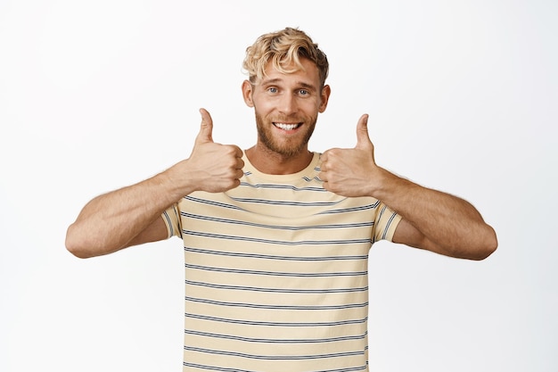 Foto knappe blonde man die lacht en zijn duimen omhoog laat zien, zoals en het ermee eens is dat hij op een witte achtergrond staat