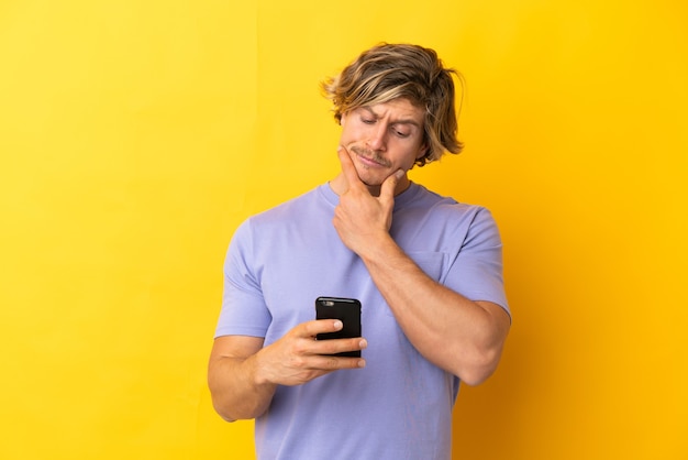 Knappe blonde man geïsoleerd op geel denken en een bericht verzenden