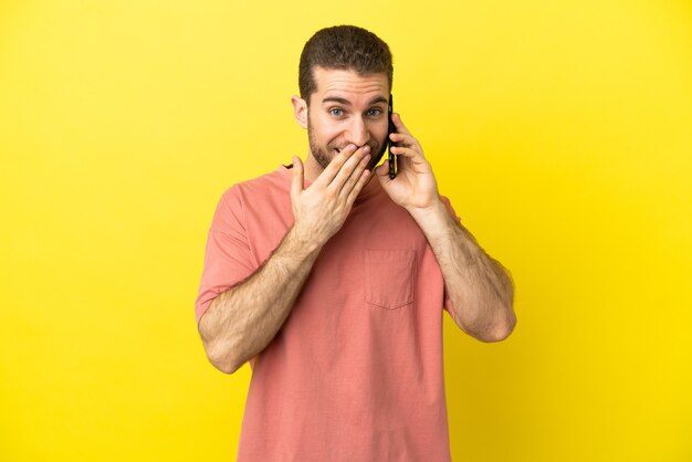 Knappe blonde man met behulp van mobiele telefoon over geïsoleerde achtergrond gelukkig en glimlachend die mond bedekken met hand