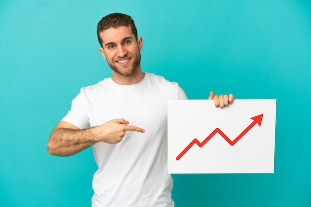 Knappe blonde man over geïsoleerde blauwe achtergrond met een bord met een groeiend statistiek pijlsymbool en erop wijzend