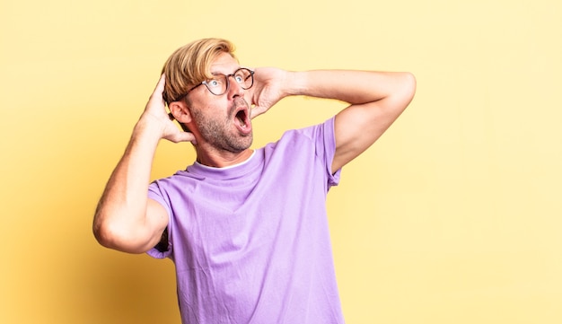 Knappe blonde volwassen man met open mond, geschokt en geschokt vanwege een vreselijke fout, handen opstekend
