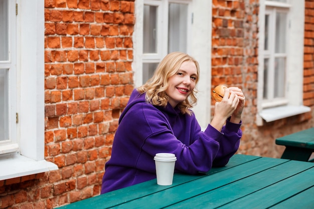 Knappe blonde vrouw eet hamburger op het terras, kopje koffie op de houten tafel.