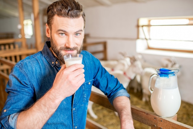 Knappe boer die verse melk proeft die in de geitenschuur staat met geiten op de achtergrond. Natuurlijke melkproductie en landbouw