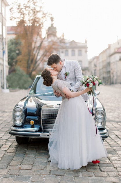 Knappe bruidegom omhelst de taille van de bruid terwijl ze haar lippen kust, staande in de buurt van de zwarte oude auto in het historische stadscentrum. Lviv, Oekraïne