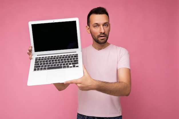 Knappe brunet man met laptopcomputer kijken camera in t-shirt op geïsoleerde roze achtergrond.