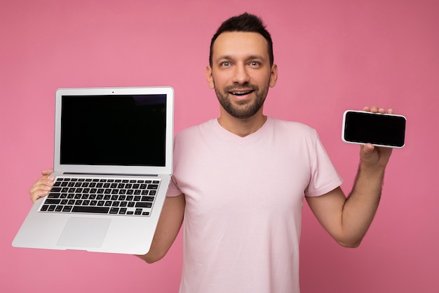 Knappe brunet ongeschoren man met laptop en mobiele telefoon kijken naar camera in t-shirt op geïsoleerde roze achtergrond.