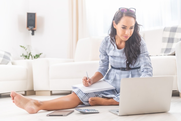 Knappe brunette in een jurk zit blootsvoets op de vloer van haar woonkamer, werkt aan een notitieboekje, schrijft notities.