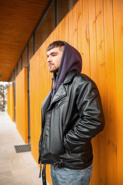 Foto knappe brutale jonge hipster-man in trendy vrijetijdskleding met een leren jas en hoodie in de buurt van een gele houten muur