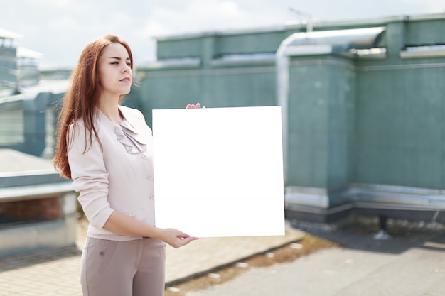 Knappe businesslady in beige pak en bruine broek staan op het dak en tonen een leeg bord