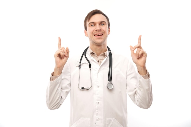 Knappe dokter in witte medische jas met stethoscoop wijst vinger naar lege ruimte geïsoleerd op een witte achtergrond met kopie ruimte. Demonstreert product, laat enthousiast iets zien