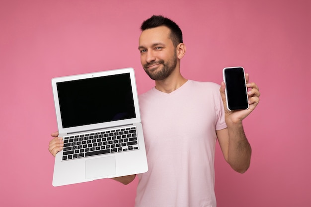 Knappe donkerbruine man met laptop en mobiele telefoon kijken naar camera in t-shirt op geïsoleerde roze achtergrond.