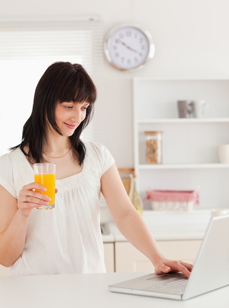 Knappe donkerbruine vrouw die een glas jus d&#39;orange houdt terwijl het ontspannen met haar laptop