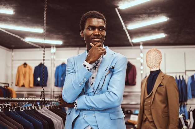 Knappe elegant geklede Afrikaanse man poseren met de hand op de kin terwijl hij in een klassieke herenkledingwinkel staat.