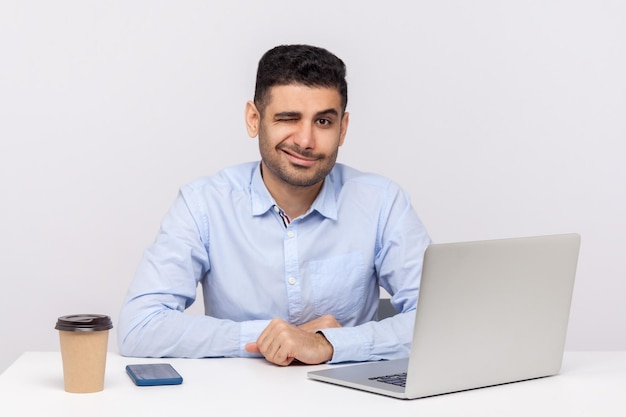 Knappe elegante zakenman zit kantoor werkplek met laptop op bureau, speels knipogend naar camera en flirten, knipperende ogen met interessant idee. studio-opname geïsoleerd op een witte achtergrond