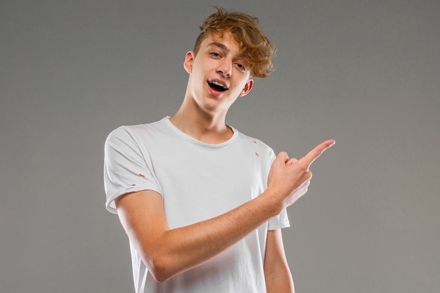 Knappe emotionele tienerjongen poseren in studio tegen grijs, man in een grijs t-shirt toont zijn vinger naar rechts