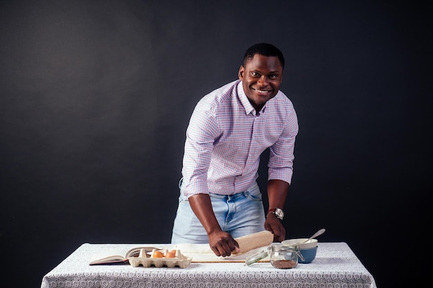 Knappe en jonge Afro-Afrikaanse man bereidt zelfgemaakte taarten American Pie van vers deeg, handen vuil door bloem op tafel zijn eieren deegroller en receptenboek op een zwarte achtergrond in de studio
