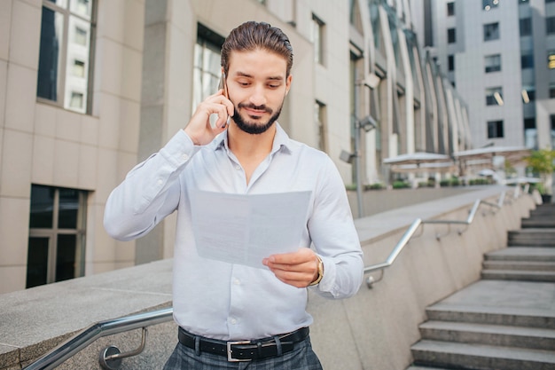 Knappe en positieve jonge man staat op stappen. hij houdt een stuk papier vast en glimlacht. zakenmanbesprekingen op telefoon. hij kijkt naar document.