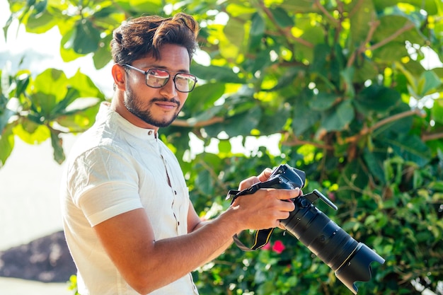 Knappe en zelfverzekerde Indiase man fotograaf met een grote professionele camera fotograferen fotograferen op het strand. fotosessie op zomervakantie op de achtergrond van groene tropische bomen