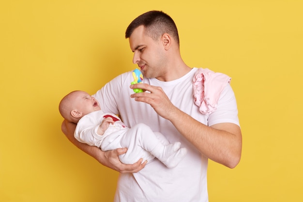 Knappe Europese man met een casual wit t-shirt staat tegen de gele muur met zijn pasgeboren baby, kijkt naar zijn kleine dochter, houdt een zitzak vast en toont aan het kind, in een poging het kind te kalmeren.