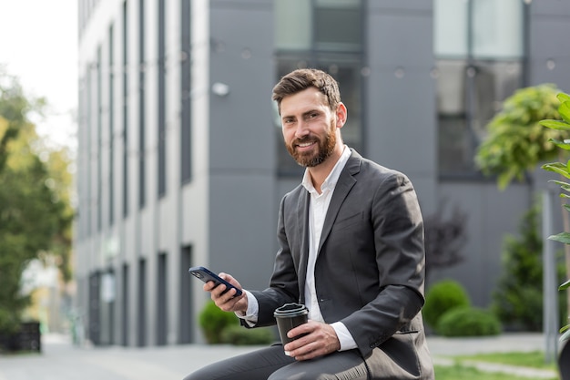 Knappe gelukkig bebaarde zakenman zittend op een bankje met kopje koffie gebruik mobiele telefoon bladeren smartphone in handen leest goed nieuws en vrolijke glimlach. Buiten een moderne stedelijke straatachtergrond. Stadspark