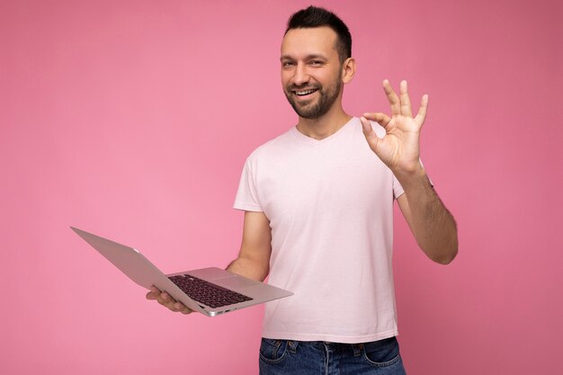 Knappe gelukkig en grappige donkerbruine man met laptopcomputer en gebaar tonen oke camera in t-shirt kijken op geïsoleerde roze achtergrond.