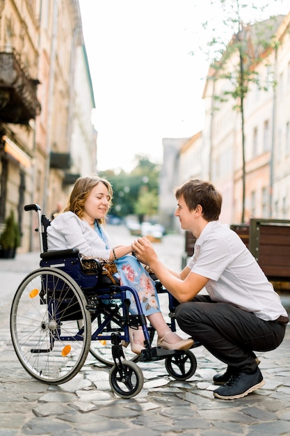 Knappe gelukkige man die en hand van zijn mooie geliefde blonde gehandicapte vrouw in rolstoel glimlachen houden terwijl het lopen samen op de straat van de oude stad