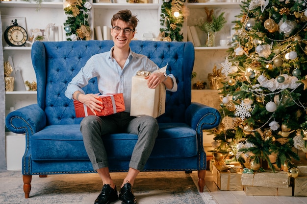 Knappe gelukkige mens met het glimlachen gezichtszitting op bank met giftboxes op zijn benen in ruimte met cristmasboom.