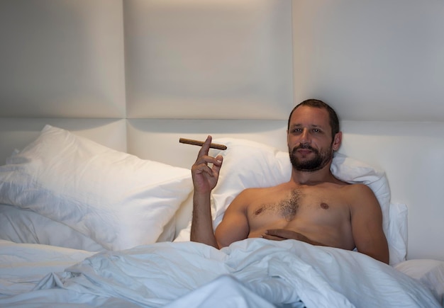 Knappe gespierde man wil sigaret roken op zijn bed naakte bebaarde blanke man liggend in bed onder een witte deken met wil cubaanse sigaar aansteken knappe rokende man in slaapkamer rust