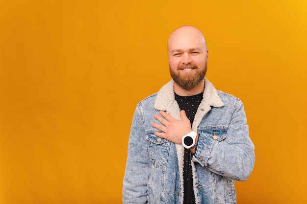 Knappe glimlachende bebaarde kale man in jeansjasje die de hand dicht bij het hart houdt