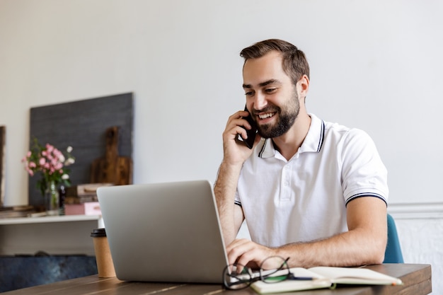 Knappe glimlachende jonge, bebaarde man die thuis aan tafel zit, laptopcomputer gebruikt, praat op mobiele telefoon