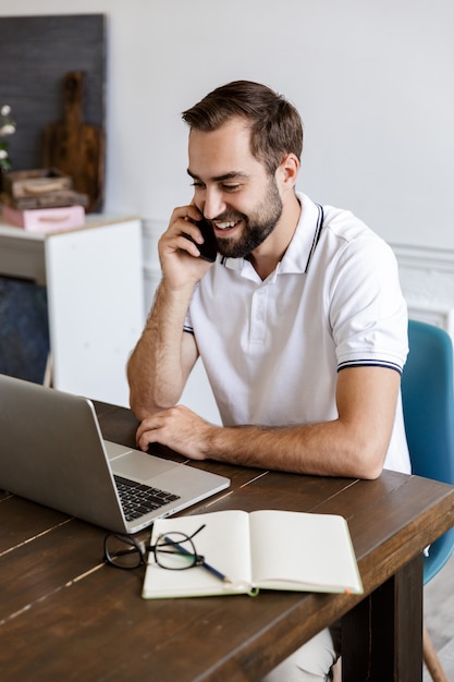 Knappe glimlachende jonge, bebaarde man die thuis aan tafel zit, laptopcomputer gebruikt, praat op mobiele telefoon
