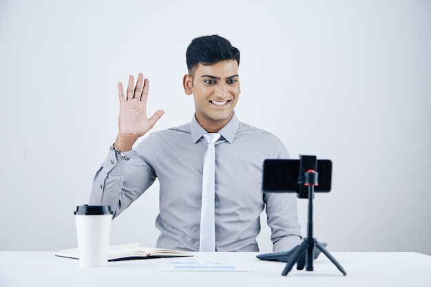 Knappe glimlachende jonge Indiase ondernemer die aan het bureau zit met een kopje koffie en video die zijn collega of zakenpartner belt