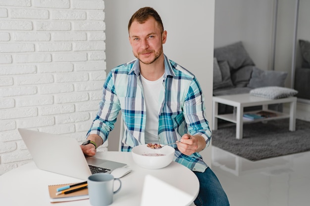 Foto knappe glimlachende man in hemd zittend in de keuken thuis aan tafel online werken op laptop